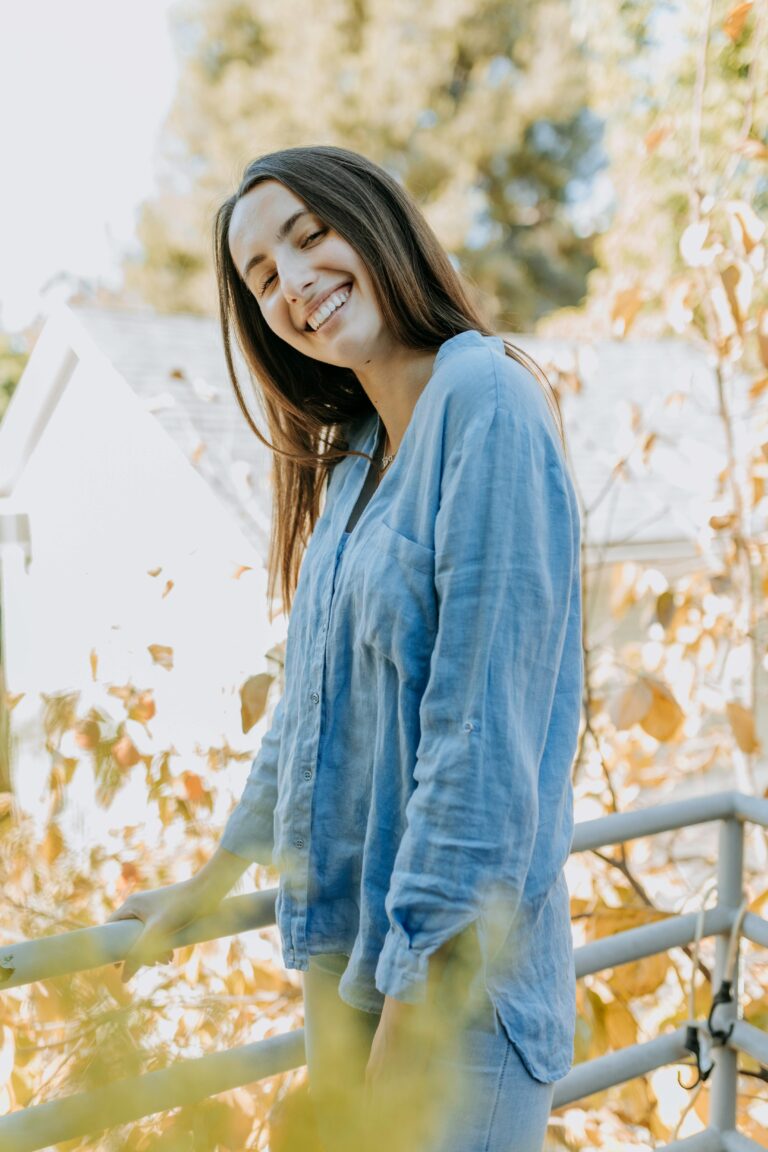 A woman in denim smiles warmly outdoors amidst autumn foliage on a sunny day.